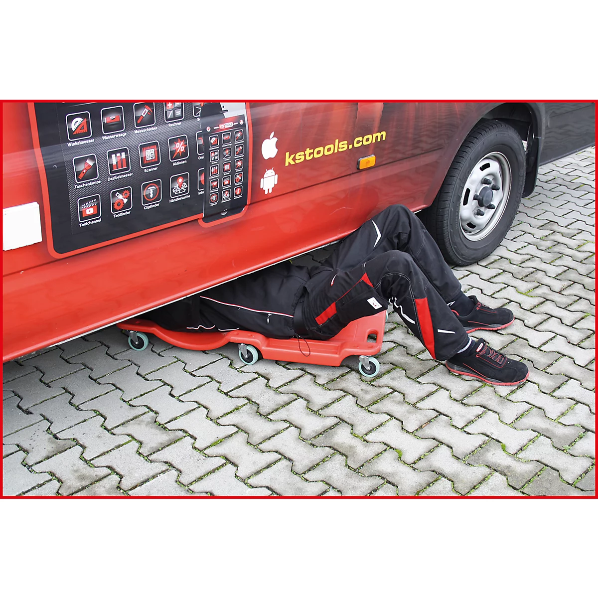 Ein Mechaniker liegt unter einem roten Fahrzeug auf einem Rollbrett, die Beine hervorschauend. Auf dem Fahrzeug steht ks-tools.com.